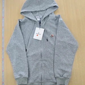 Grå zip-hoodie från Moncler - Grå hoodie med dragkedja från Moncler. Klassisk modell med huva, ribbade muddar och logotyp på bröstet. Perfekt för en avslappnad stil och enkel att matcha med andra plagg.