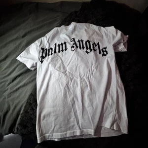 Vit t-shirt från Palm Angels - Säljer en vit t-shirt från Palm Angels med stor svart logotyp på ryggen och mindre logga tryckt vid halsen framtill. Klassisk passform och rund halsringning. Perfekt för dig som gillar streetwear och vill sticka ut med en ikonisk design.