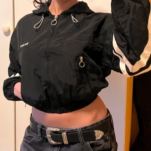 Svart croppad vindjacka med huva från Bershka - Säljer en svart croppad vindjacka från Bershka med huva och dragkedja. Går att justera på olika ställen. Perfekt för en sportig och trendig look.