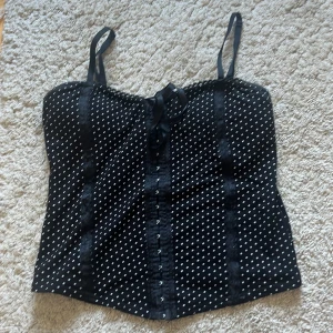 Svart prickig korsettopp från New Look - Säljer en svart korsettopp från New Look med vita prickar och smala axelband. Toppen har dekorativa knappar framtill, spetsdetaljer och en liten rosett vid bysten. Perfekt för att lyfta vilken outfit som helst! Pris kan diskuteras!