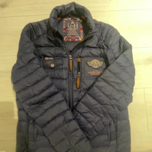  Pufferjacka Morris - Säljer en blå pufferjacka från Morris med broderad logga på bröstet och snygga detaljer i mockaimitation vid dragkedjorna. Jackan har hög krage, lång ärm och flera fickor framtill. Perfekt för kyliga dagar!❤️👍🏼