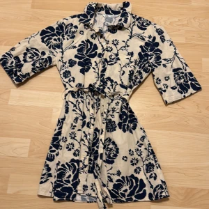 Blommig skjortklänning med knytskärp från Zara - Säljer en beige skjortklänning från Zara med stora marinblå blommor. Klänningen har krage, knappar framtill, trekvartsärm och ett matchande knytskärp i midjan. Perfekt för dig som gillar en avslappnad men stilren look.