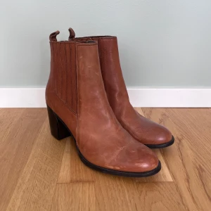 Bruna chelsea boots med klack - Säljer ett par snygga bruna chelsea boots i skinn med hög klack och rund tå. Resår på sidorna och dragkedja på insidan gör dem enkla att ta på. Perfekta för dig som vill ha en stilren och tidlös look.