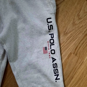 Grå mjukisbyxor från U.S. Polo Assn. - Snygga ljusgrå mjukisbyxor från U.S. Polo Assn. med tryckt logga längs benet och en bakficka med liten flaggdetalj. Perfekta för en avslappnad stil och sköna att ha på sig hela dagen.