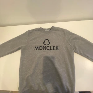 Grå sweatshirt från Moncler - Säljer en stilren grå sweatshirt från Moncler med svart logotyp och text på bröstet. Tröjan har rund halsringning och långa ärmar. Perfekt för en avslappnad och trendig look.