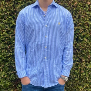 Blåvit randig skjorta från Polo Ralph Lauren - Säljer en klassisk blå och vit randig skjorta från Polo Ralph Lauren, modell Stanton Custom Fit. Skjortan har långa ärmar, knappar framtill och den ikoniska broderade loggan i gult på bröstet. Perfekt för en stilren look!
