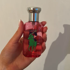 Rosa parfymflaska från Ralph Lauren Big Pony 2 - Säljer en rosa parfym från Ralph Lauren Big Pony Collection nummer 2. Flaskan är genomskinlig med silverfärgad kork, blå siffra 2 på ena sidan och grön hästlogga på andra. Perfekt för dig som gillar fräscha och sportiga dofter.