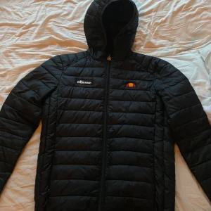Svart pufferjacka från Ellesse med huva - Säljer en svart pufferjacka från Ellesse med huva och dragkedja. Jackan har quiltad design och Ellesse-logga på bröstet. Perfekt för kyligare dagar och enkel att matcha med olika outfits.