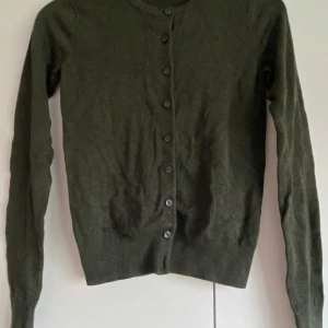 Mörkgrön kofta från H&M - Säljer en enkel och stilren mörkgrön kofta från H&M. Koftan har långa ärmar, rund halsringning och knäppning framtill med matchande knappar. Perfekt att slänga över en topp eller klänning för en snygg lager-på-lager-look.