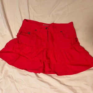 Vintage rosa från Chunky - Säljer en rosa kortkjol från Chunky med volangdetaljer och fickor framtill. Kjolen har en klassisk jeanskjolsdesign med knapp och dragkedja i midjan. Perfekt för dig som vill ha en färgstark och lekfull stil.