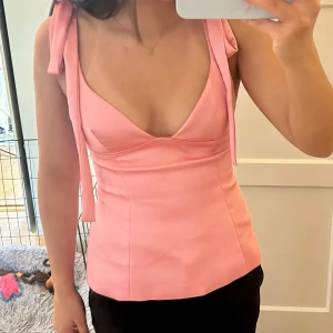 Rosa linnetopp med knytband - Säljer en söt rosa linnetopp med djup v-ringning och knytband på axlarna. Toppen har figurnära passform och är tillverkad i ett slätt material. Perfekt för varma dagar eller när du vill känna dig extra fin.