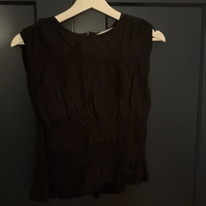Svart ärmlös blus med rynk - Säljer en svart blus med rund halsringning och rynkade detaljer. Toppen är ärmlös och har en lätt volang nertill för en snygg touch. Perfekt att styla med jeans eller kjol för en clean look.