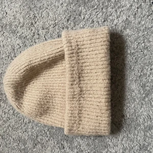 Beige ribbad mössa från Gina Tricot - Mysig ribbstickad mössa i beige från Gina Tricot. Klassisk modell med uppvikt kant, perfekt för kyliga dagar. Enkel och stilren design som passar till det mesta. Tillverkad i mjukt material för extra komfort.