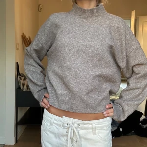 Beige stickad polotröja - Mysig beige stickad polotröja med ribbad hög krage och muddar. Tröjan har en loose passform och långa ärmar, perfekt för kyliga dagar. Enkel att matcha med både jeans och kjol för en avslappnad look.