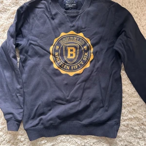 Mörkblå sweatshirt från Bondelid - Mörkblå sweatshirt från Bondelid med stort gult tryck framtill och klassisk rund hals. Tröjan är tillverkad i 100% bomull och har långa ärmar samt ribbade muddar vid ärmslut och nederkant. Perfekt för en avslappnad och sportig stil. Stl M