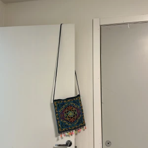 Broderad axelväska med tofsar - Färgglad axelväska med broderat mönster i blått, gult, rött och grönt. Väskan har fyrkantig form, lång smal axelrem och små tofsar i olika färger längs nederkanten. Perfekt för dig som gillar unika accessoarer med boho-vibe.