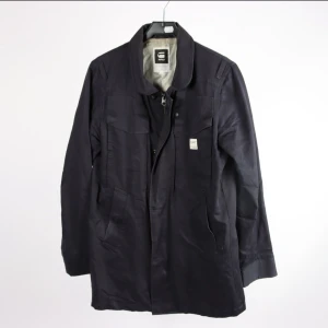 Bkå jacka från G-Star RAW, stl M - Stilren Blå jacka från G-Star RAW i storlek M. Jackan har klassisk krage, flera fickor framtill och stängs med dragkedja och knappar. Materialet är en mix av bomull och linne med polyesterfoder, vilket ger en skön känsla och snygg passform.