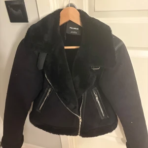 Svart pälsjacka från Pull&Bear - Säljer en svart pälsjacka från Pull&Bear med stora slag, dragkedjor och mjukt foder. Jackan har en croppad passform och coola detaljer som synliga blixtlås på fickorna. Perfekt för dig som vill ha en snygg och varm look i vinter.
