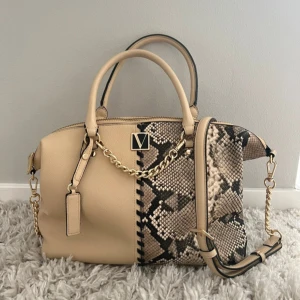 Beige handväska från victoria secret med kedja - Snygg beige handväska från Victoria Secret med coolt ormmönster på ena sidan och guldfärgad kedjedetalj. Väskan har både handtag och axelrem, dragkedja upptill och detaljer i skinnimitation. Perfekt för dig som vill sticka ut med en trendig accessoar.