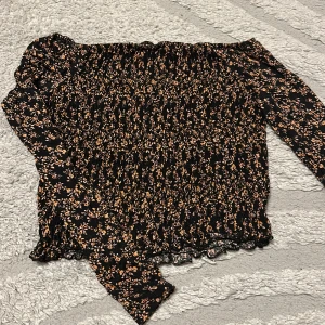 Svart blommig offshoulder topp - Supersöt svart offshoulder topp med små blommor i beige och rosa toner. Smockad över hela kroppen för en tight passform och långa ärmar. Perfekt för dig som gillar romantisk och trendig stil.