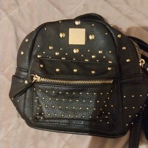 Svart ryggsäck med guldnitar - Cool svart ryggsäck i skinnimitation med massor av guldiga nitar och detaljer. Väskan har en framficka med dragkedja och justerbara axelband. Perfekt för dig som vill sticka ut med en edgy accessoar.