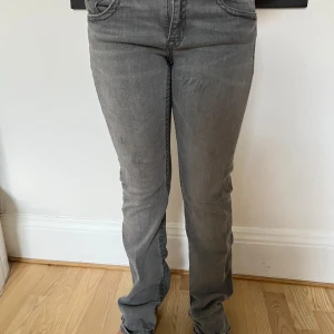 Grå bootcut jeans med fickdetaljer - Snygga grå jeans med bootcut passform och klassiska fem fickor. Bakfickorna har lock och knappdetalj för extra stil. Jeansen är i ett mjukt bomullsmaterial med en lätt tvättad look. Perfekta för dig som gillar en avslappnad men trendig vibe.