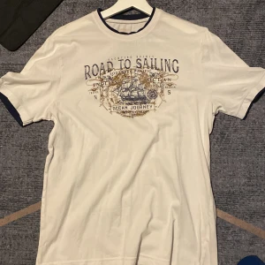 Vit t-shirt LC Waikiki med tryck - Snygg vit t-shirt från LC Waikiki med marinblå detaljer vid krage och ärmslut. Framsidan har ett stort tryck med texten 'Road to Sailing' och motiv av ett skepp. Perfekt för dig som gillar nautisk stil och vill sticka ut lite extra.