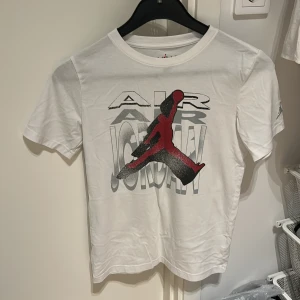 Vit Air Jordan t-shirt - Vit t-shirt från Jordan med stort Air Jordan-tryck framtill och ikonisk röd Jumpman-logga. Klassisk passform med korta ärmar och rund hals. Materialet är en mix av bomull och polyester, vilket gör den både mjuk och skön att bära.