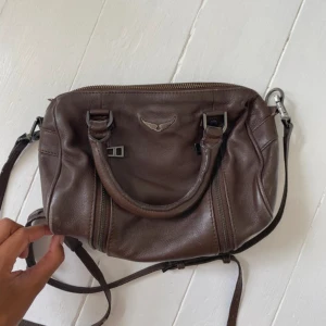 Brun skinnväska Zadig & Voltaire - Snygg brun handväska från Zadig & Voltaire i äkta skinn. Väskan har både handtag och justerbar axelrem, samt coola dragkedjedetaljer på sidorna. Perfekt storlek för det viktigaste och med klassisk logga framtill.