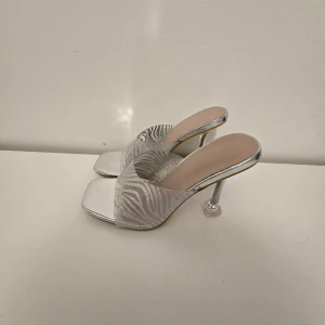 Silvriga klackar med zebramönster - Glammiga silvriga klackar med transparent zebramönstrad rem och fyrkantig tå. Snygg, hög klack och öppen tå ger en trendig vibe. Perfekta för dig som vill sticka ut med metallic och coolt mönster. Materialet är syntet och skon har en plattformslook.