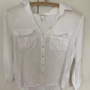 Vit linneskjorta med fickor från H&M - Vit skjorta i linne från H&M med klassisk krage, två bröstfickor med lock och knäppning framtill. Skjortan har lång ärm och en avslappnad passform, perfekt till jeans eller shorts. Enkel och stilren design som funkar året runt.