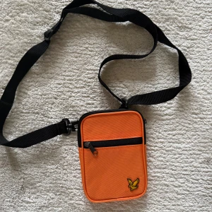 Orange axelväska från Lyle & Scott - Snygg orange axelväska från Lyle & Scott med svart justerbar rem och dragkedja framtill. Väskan har en broderad gul logga med örn på framsidan och är tillverkad i slitstark polyester. Perfekt för att bära det viktigaste när du är på språng.