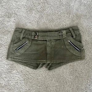 Olivgröna lågmidjade shorts med dragkedjor - Snygga olivgröna shorts i lågmidjad modell i storlek xs/s med coola dragkedjor på fickorna framtill och dekorativa knappar. Shortsen har bälteshällor och två fickor med lock och knapp bak. Perfekta för en chill och trendig sommarlook. Midjemått: ca 70cm💞