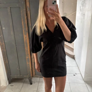 Svart kort klänning med puffärm - Svart kort klänning med v-ringning och puffiga trekvartsärmar. Enkel och stilren design som ger en cool vibe. Klänningen har en rak passform och är tillverkad i ett lätt tyg, perfekt för varmare dagar.