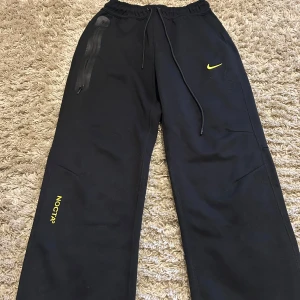 Svarta NOCTA x Nike mjukisbyxor - Svarta mjukisbyxor från NOCTA x Nike med gul logga och detaljer. Byxorna har elastisk midja med snörning, raka ben och en vattentät dragkedja på ena fickan. Tillverkade i mjuk bomullsmix, perfekt för chill eller streetwear.