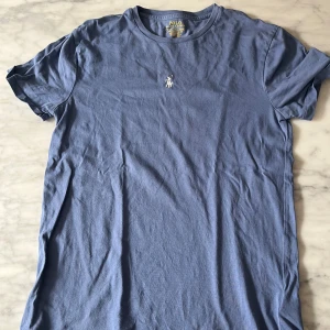 Blå t-shirt från Polo Ralph Lauren - Snygg blå t-shirt från Polo Ralph Lauren med klassisk rund hals och broderad logga på bröstet. Modellen är slim fit och har korta ärmar. Tillverkad i mjuk bomull som känns skön mot huden. Perfekt för en clean och stilren look.