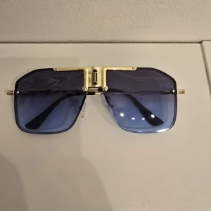 Blåtonade solglasögon med gulddetaljer - Stilrena solglasögon med stora, fyrkantiga blåtonade glas och en unik guldfärgad metallbåge. Snygg detalj över näsryggen och tunna skalmar. Perfekta för dig som vill sticka ut med en modern och lyxig look.