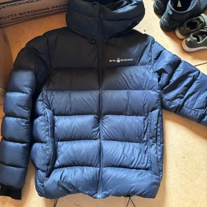 Blå pufferjacka från Sail Racing XL - Säljer en blå pufferjacka från Sail Racing. Jackan är i storlek XL men passar mig som brukar ha M,så det beror på hur tight man vill att den ska sitta. Jackan har en hög krage, huva och dragkedja framtill. Den är vadderad och har en clean look med logga på bröstet. Perfekt för kalla dagar när du vill hålla stilen och värmen.