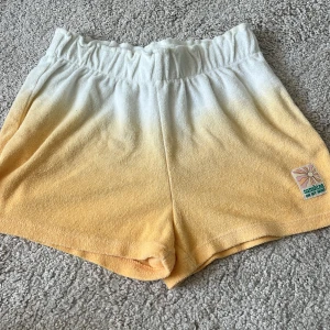 Gula och vita frottéshorts Sunshine - Mjuka shorts i frotté med snygg gul till vit ombre-effekt och resår i midjan. Liten patch med solmotiv och texten 'sunshine on my mind' på benet. Perfekta för chill dagar eller strandhäng. Aldrig använda näst in på