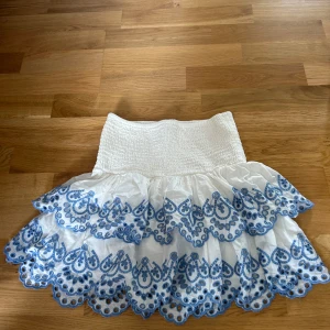Vit volangkjol med blå broderi Lindex - Supersöt vit kortkjol från Lindex med bred smock i midjan och två lager volanger. Kjolen har blå broderade detaljer längs volangkanterna som ger en riktigt somrig vibe. Perfekt för dig som gillar en lekfull och fräsch stil.