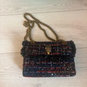 Axelväska i tweed med kedja - Snygg axelväska i mörk tweed med inslag av rött, blått och svart. Väskan har en grov guldfärgad kedja och ett dekorativt spänne framtill. Perfekt för att lyfta vilken outfit som helst med sin unika textur och coola detaljer. Ny skick 