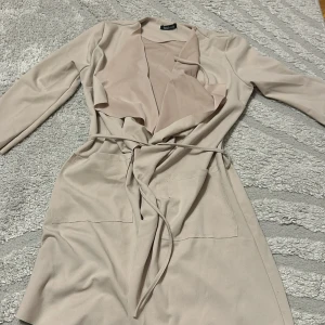 Beige trenchcoat från Boohoo - Snygg beige trenchcoat från Boohoo med långa ärmar, öppen front och bälte i midjan. Jackan har stora fickor framtill och ett mjukt, lätt material som ger en avslappnad men stilren look. Perfekt för dig som gillar minimalistisk stil och neutrala färger.
