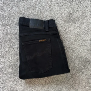 Svarta jeans från Nudie Jeans, stl 30/30 - Svarta jeans från Nudie Jeans med klassisk femficksdesign och diskret logga på bakfickan. Jeansen har normal passform och är tillverkade i mjuk bomull med lite stretch för extra komfort. Perfekta för dig som gillar stilrena och enkla svarta jeans.