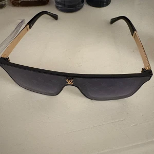 Louis Vuitton svarta solglasögon - Säljer ett par feta svarta Louis Vuitton solglasögon med guldiga detaljer och LV-logga framtill. Glasen är mörka med diskret monogrammönster och bågarna har snygga guldinslag med rutigt mönster. Perfekt accessoar för att sticka ut.