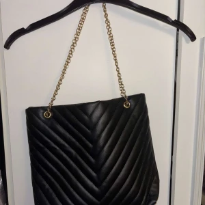Svart quiltad axelväska med kedja - Snygg svart axelväska med quiltad framsida och guldfärgad kedjerem. Väskan har en mjuk struktur och dragkedja upptill.