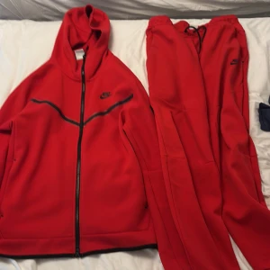 Röd Nike träningsdress med huva - Nike träningsset i rött med svart logga. Hoodien har dragkedja, huva och svarta detaljer över bröstet. Byxorna har resår och snörning i midjan. Perfekt för dig som gillar sportig och bekväm stil.