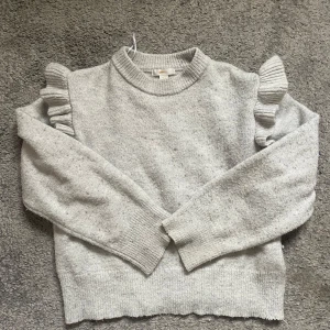 Grå stickad tröja med volang Gina Tricot - Superfin ljusgrå stickad tröja från Gina Tricot med volangdetaljer på axlarna. Tröjan har rund halsringning, långa ärmar och ribbade muddar. Perfekt för dig som gillar en söt och trendig look!