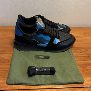 Valentino Garavani Rockrunner sneakers - Snygga Valentino Garavani Rockrunner sneakers i svart och blått med coolt kamouflagemönster. Skorna har svarta nitar på sulan, snörning och detaljer i skinn och mocka. Perfekta för dig som vill sticka ut med exklusiva och trendiga sneakers.