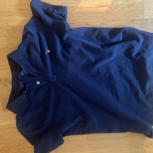 Marinblå pikétröja från Gant - Snygg marinblå pikétröja från Gant med klassisk krage och två knappar framtill. Tröjan har Gant-logga broderad på bröstet och är tillverkad i mjuk bomull. Perfekt för en stilren och avslappnad look. Passar storlek S