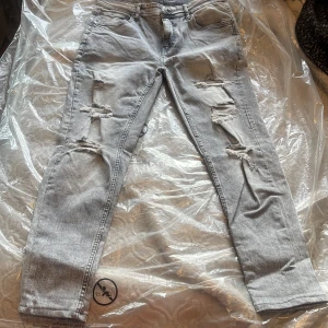 Ljusgrå ripped jeans från Black Squad - Säljer ett par ljusgrå jeans från Black Squad med snygga slitningar och hål på båda benen. Jeansen har klassisk femficksdesign, dragkedjegylf och raka ben. Perfekta för dig som gillar en edgy streetstil.